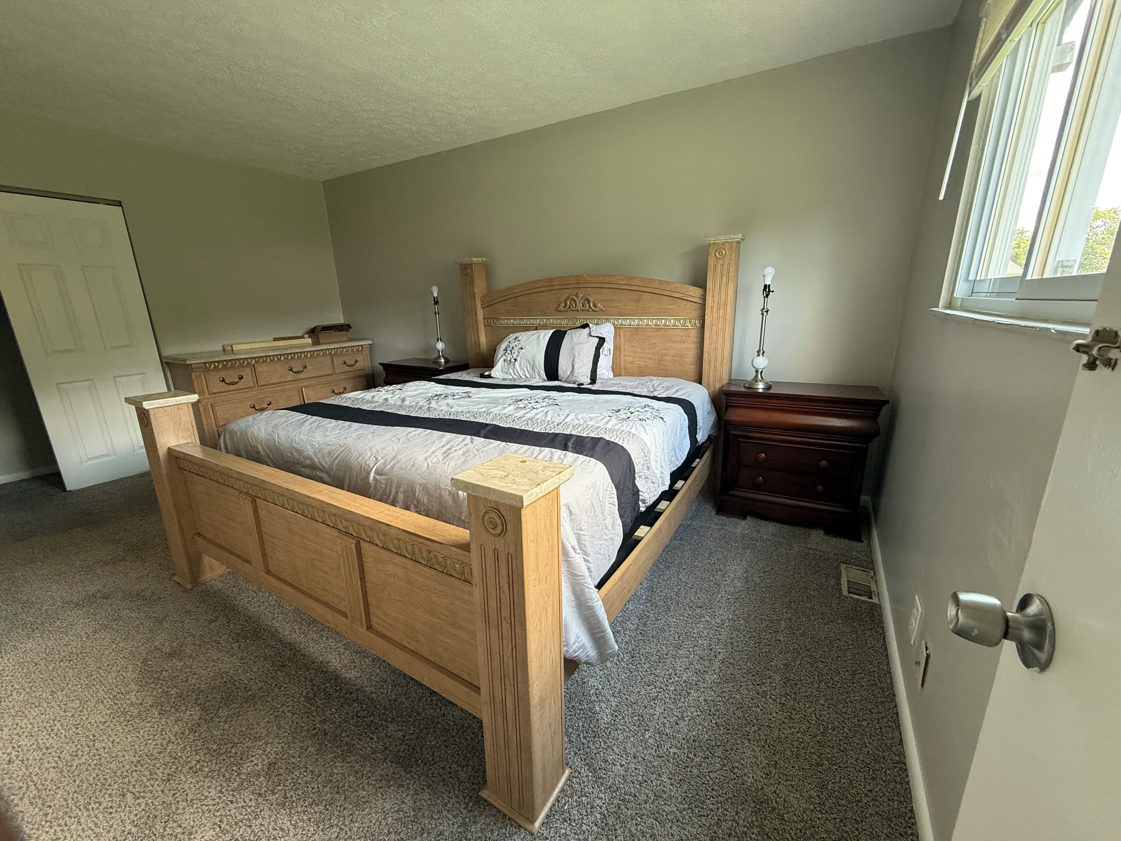 Quarto com cama de madeira, mesa de cabeceira, cômoda e porta.