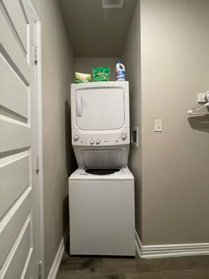 A laundry room with a stacked washer and dryer, a door, and a wall-mounted light switch. - Cozy 1BR Cottage Retreat Ideal for Extended Stays (Hurst)