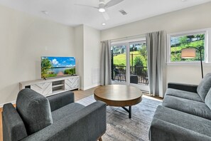 A modern living room with a flat-screen TV, grey sofas, a wooden coffee table, and a balcony with a view of greenery and buildings.