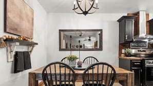 Un comedor con mesa y sillas de madera, una araña, un espejo y una cocina con electrodomésticos modernos.