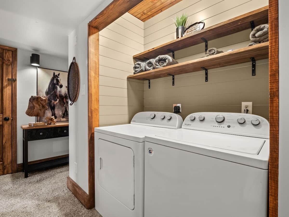A laundry room with two stacked washers and dryers, wooden shelves above, and a coat rack with a horse painting.