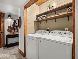 A laundry room with two stacked washers and dryers, wooden shelves above, and a coat rack with a horse painting.