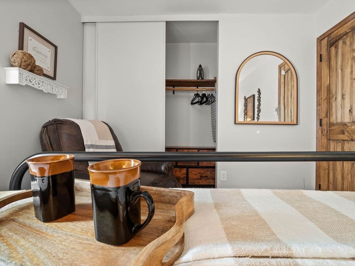Quarto de hotel com uma cama, uma mesinha de madeira com duas canecas, um cabideiro, um espelho e um guarda-roupa.