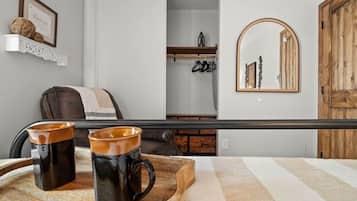Quarto de hotel com uma cama, uma mesinha de madeira com duas canecas, um cabideiro, um espelho e um guarda-roupa.