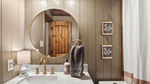 Un baño con lavamanos de mármol, grifos dorados, un espejo redondo, paneles de pared de madera y una cortina de ducha blanca.