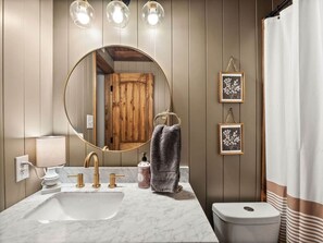 A bathroom with a marble sink, gold faucets, a round mirror, wooden panel walls, and a white shower curtain. - Fort Polk Cozy 2BR Stay l Monthly & Extended Stays (Leesville)