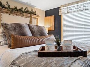 A bed with two pillows, a tray with two coffee mugs, and a window with blinds.