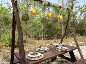 A wooden picnic table set up outdoors with plates and cutlery, surrounded by trees and hanging spherical lanterns.