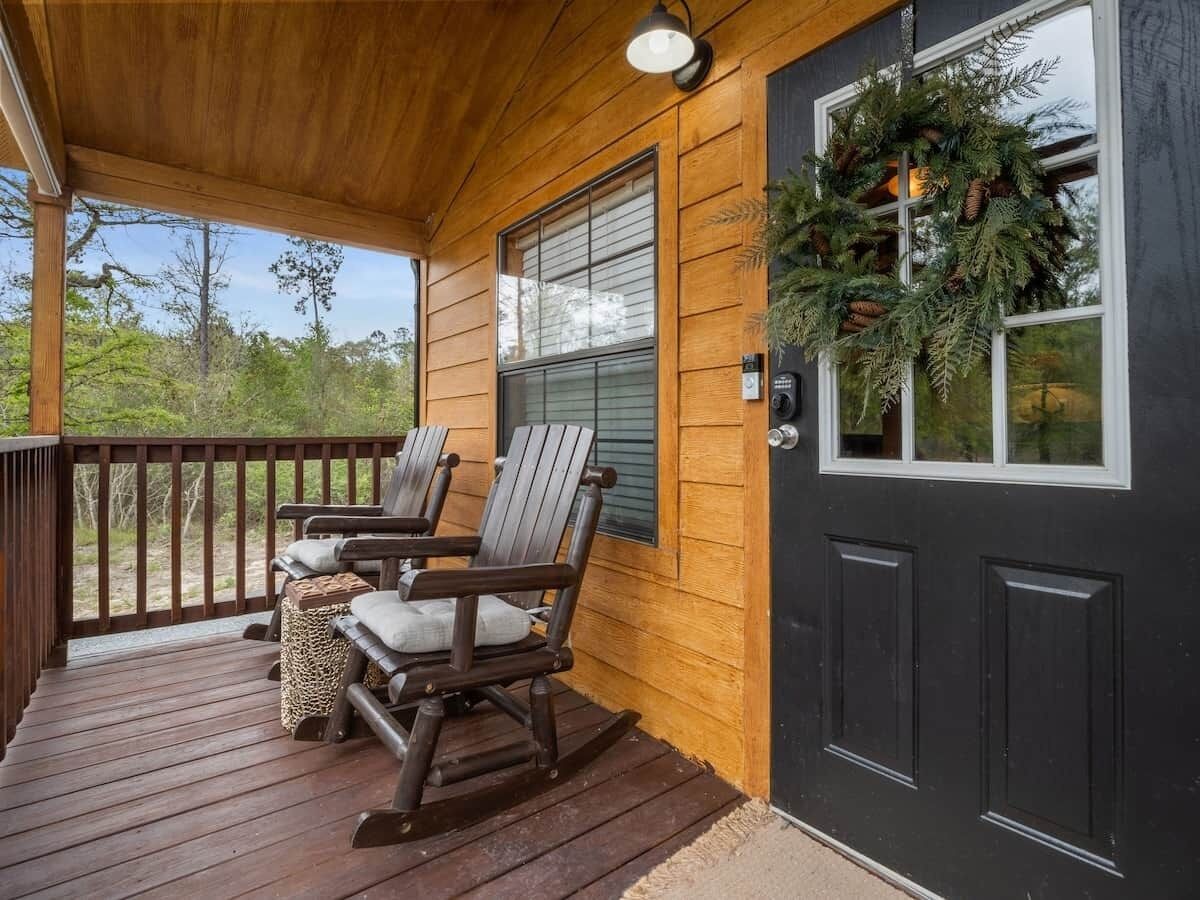 Un porche de madera con dos sillas Adirondack y una mesa pequeña, una puerta negra con una guirnalda, y vistas a árboles.