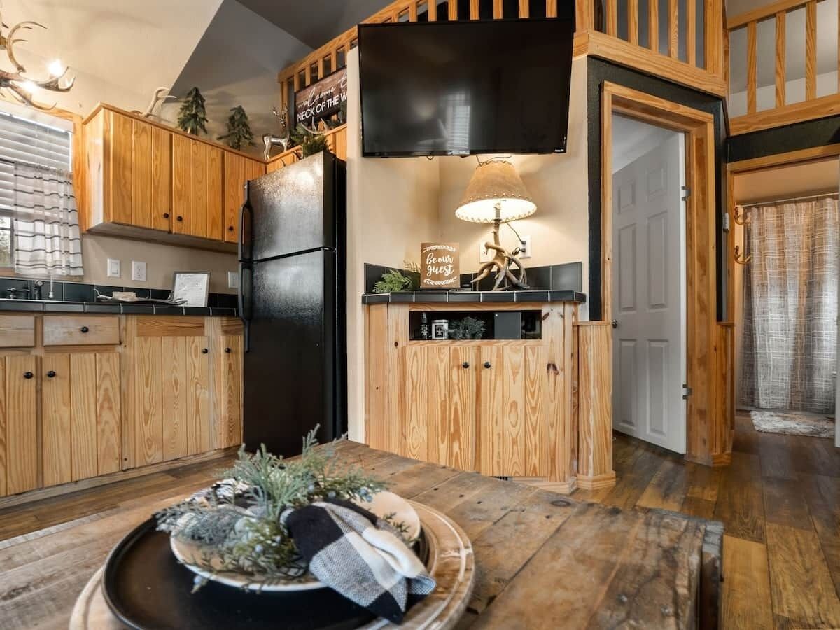 A kitchen with wooden cabinets, a black refrigerator, a dining table with a decorative centerpiece, and a wall-mounted television.