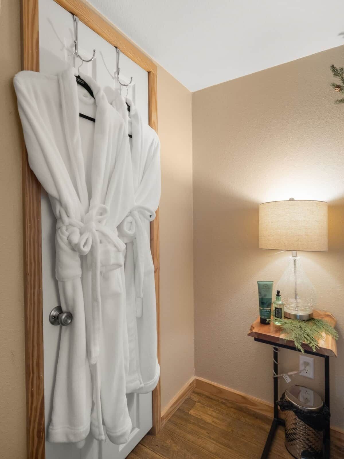 Un baño con puerta de madera, dos albornoces blancos colgados en ganchos, una lámpara de pie y una mesita con artículos de aseo.