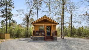 A small wooden cabin with a porch, surrounded by gravel and trees.