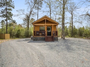A small wooden cabin with a porch, surrounded by gravel and trees.