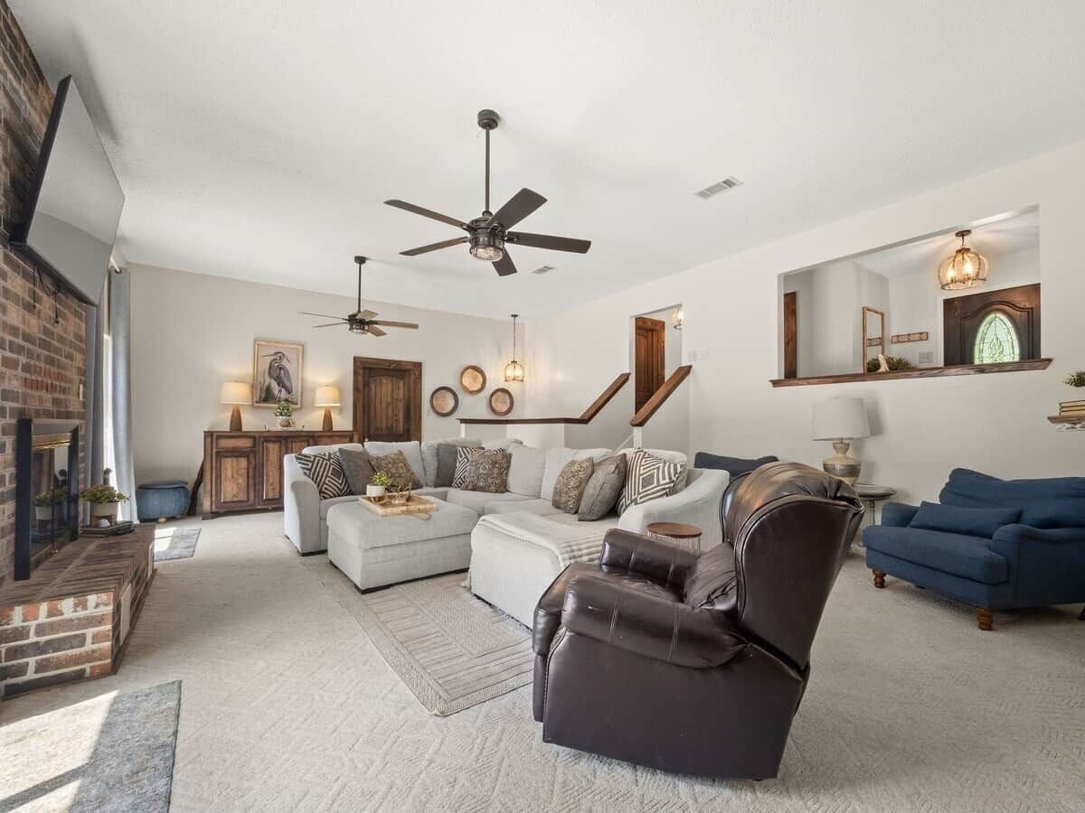 A spacious living room with a sectional sofa, a leather armchair, and a blue couch. There is a fireplace, a ceiling fan, and a staircase leading to another level.
