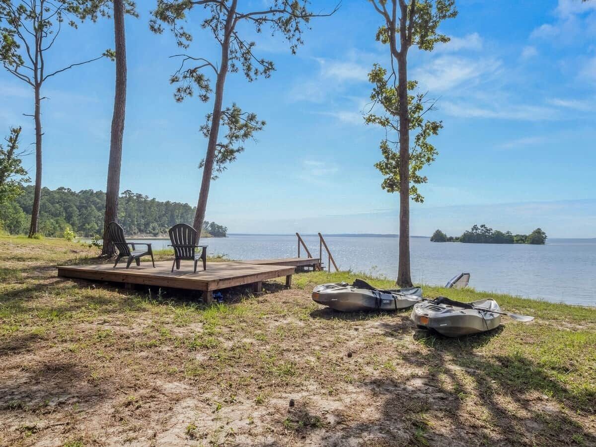 A lakeside deck with chairs and a table, and two kayaks on the ground.