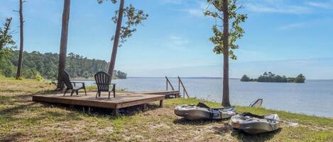 A lakeside deck with chairs and a table, and two kayaks on the ground.