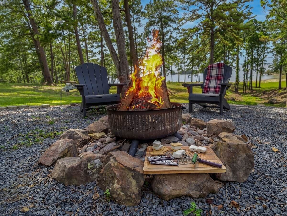 A campfire with two chairs, a fire pit, and a board with camping supplies.