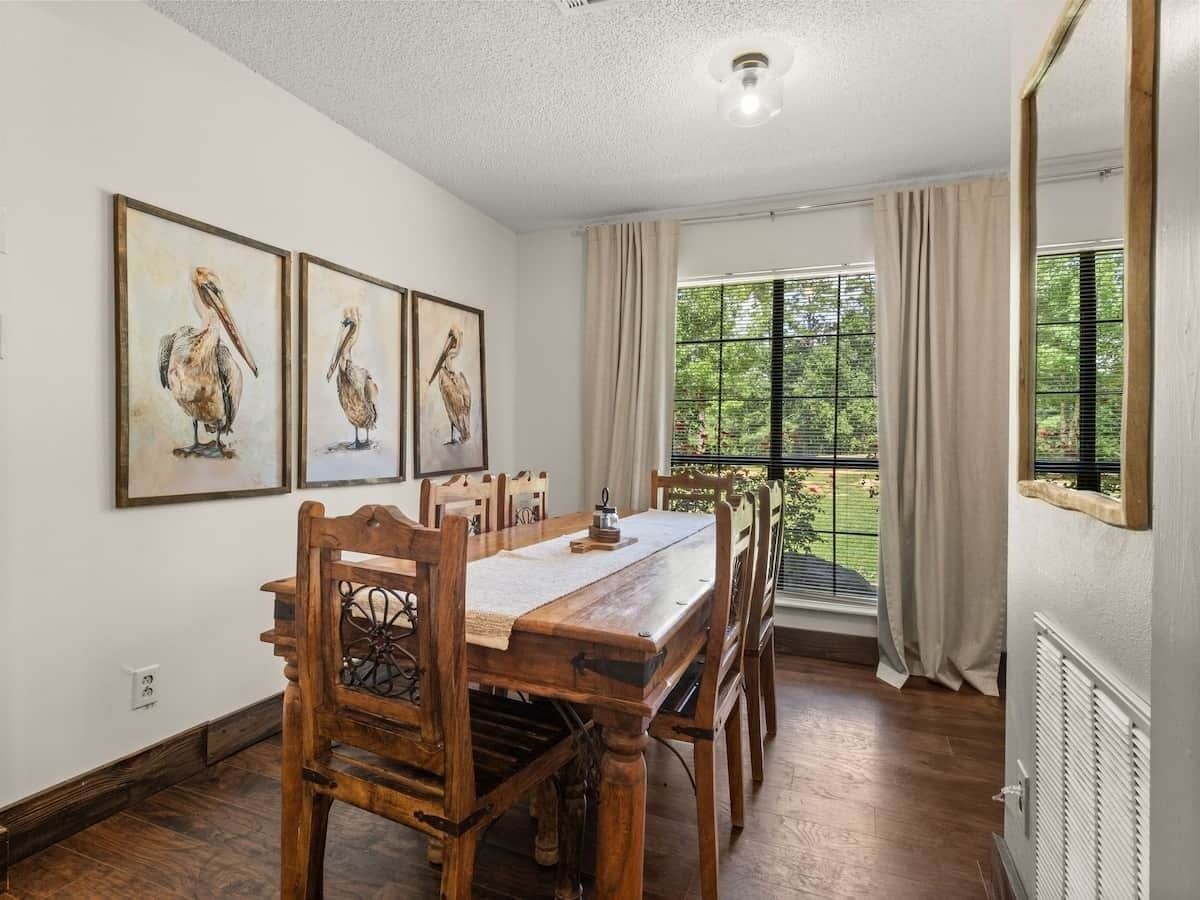 A dining room with a wooden table and chairs, framed artwork on the wall, and a window with curtains.