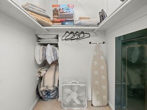 Ein kleiner Abstellraum mit Regalen, Kleidung, einem Ventilator und einem gefalteten Handtuch.