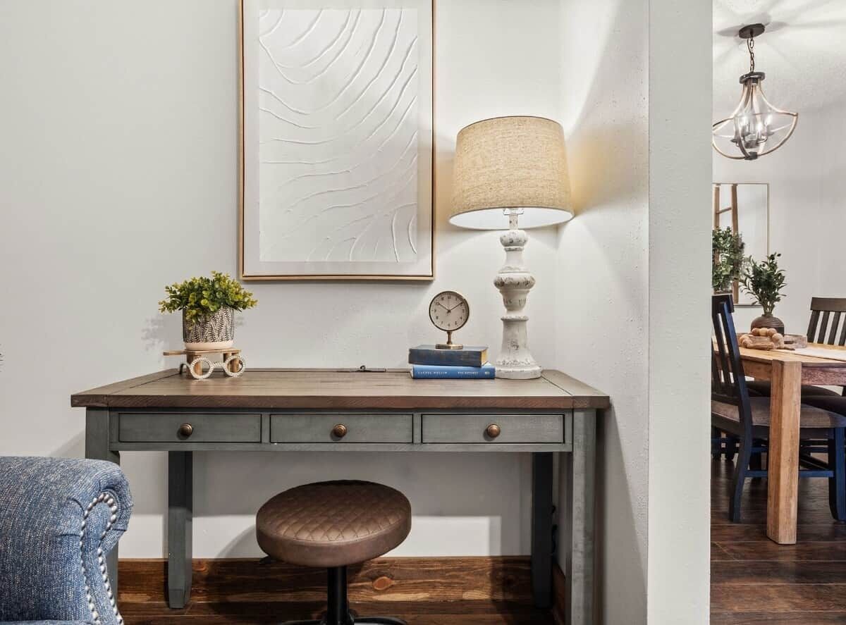 Ein Zimmer mit Schreibtisch, Stuhl, Lampe, einer Uhr und einem gerahmten Kunstwerk an der Wand.