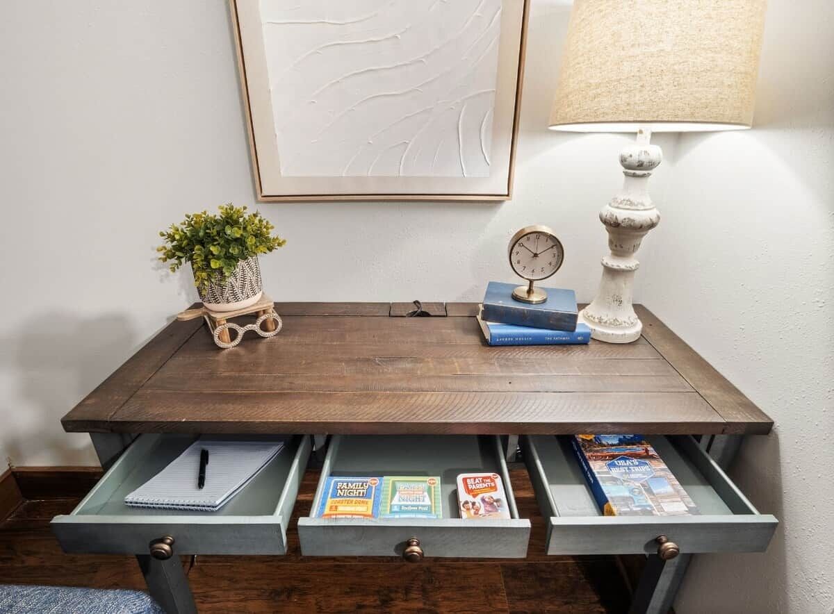 Een houten bureau met een lamp, een klok en een bloempot. Op het bureau liggen boeken en tijdschriften.