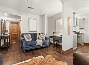 A living room with a blue sofa, wooden flooring, a desk, and a kitchen area.