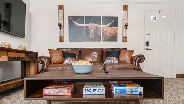 A living room with a brown leather sofa, a coffee table with a bowl of popcorn, and game boxes. - Fort Polk Cozy 2BR Stay l Monthly & Extended Stays (Leesville)