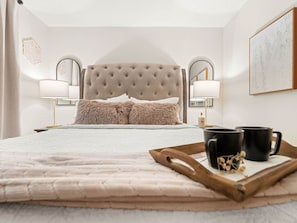 Un dormitorio con cama, lámparas de noche, una bandeja con tazas y un cuadro en la pared.