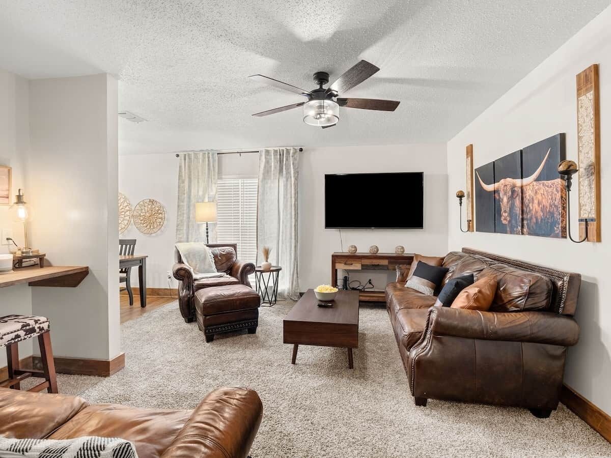 Ein Wohnzimmer mit Deckenventilator, Flachbildfernseher, Ledersofas, Couchtisch und Kunstwerken an den Wänden.