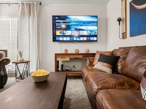 A living room with a leather sofa, a wooden coffee table with a bowl of popcorn, a TV mounted on the wall, and a small side table with decorative items.