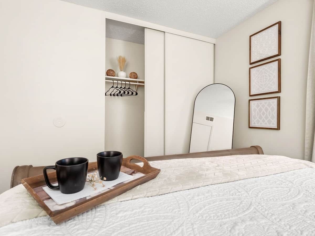 Une chambre à coucher avec un lit, un plateau en bois sur lequel reposent deux tasses noires, un mur blanc, une armoire et un miroir.