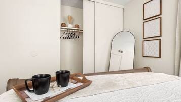Une chambre à coucher avec un lit, un plateau en bois sur lequel reposent deux tasses noires, un mur blanc, une armoire et un miroir.
