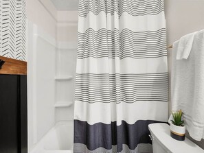 Baño con cortina de ducha a rayas blancas y negras, una bañera blanca, una toalla blanca colgada en un perchero y una pequeña planta en maceta en el borde de la bañera.
