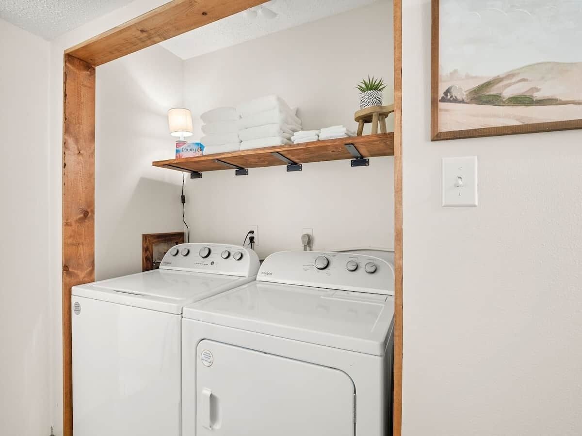 A laundry area with two white washing machines, a wooden shelf with towels and a small plant, and a painting on the wall.