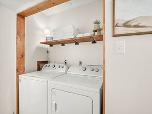 A laundry area with two white washing machines, a wooden shelf with towels and a small plant, and a painting on the wall.
