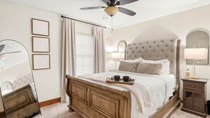 A bedroom with a large bed, a ceiling fan, a bedside table with a lamp, and a mirror. - Fort Polk Cozy 2BR Stay l Monthly & Extended Stays (Leesville)