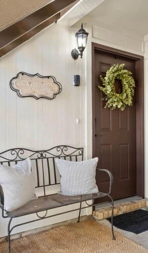 A bench with two pillows, a welcome sign, a wall-mounted light, and a wreath on a door.