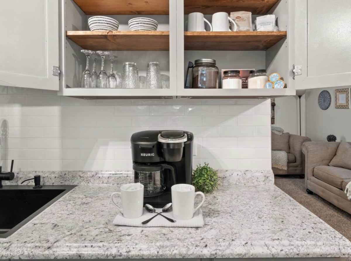Une cuisine équipée d’une machine à café intégrée, d’un plan de travail sur lequel reposent deux tasses blanches, et d’une étagère en bois au-dessus, où sont rangés divers ustensiles de cuisine.