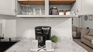 Une cuisine équipée d’une machine à café intégrée, d’un plan de travail sur lequel reposent deux tasses blanches, et d’une étagère en bois au-dessus, où sont rangés divers ustensiles de cuisine.