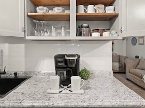A kitchen with a built-in coffee maker, a countertop with two white mugs, and a wooden shelf above holding various kitchen items.