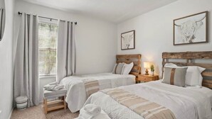 Quarto com duas camas, uma janela com cortinas, mesa de cabeceira com abajur e uma obra de arte na parede.