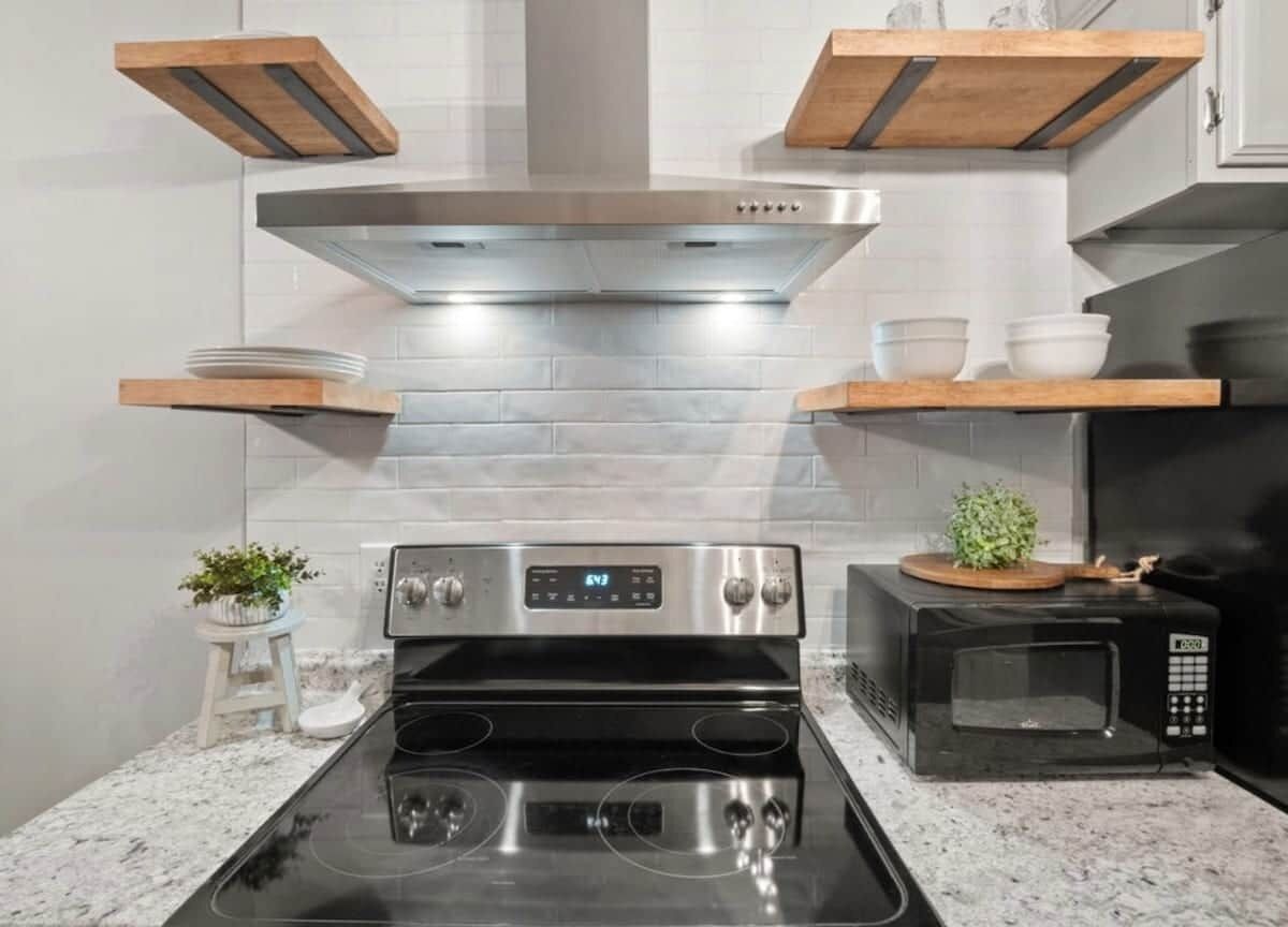 Una cocina con una campana extractora de acero inoxidable, una estufa negra, un microondas y repisas de madera sobre la encimera.