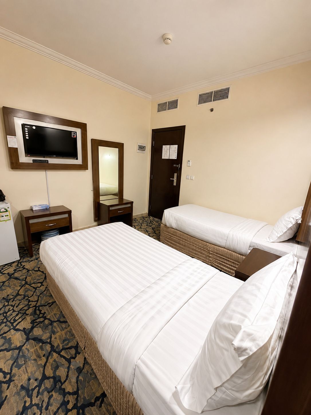 Habitación de hotel con dos camas, televisor, espejo, puerta con cerradura y alfombra estampada.