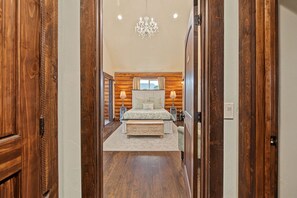 A bedroom with a large bed, bedside tables, a chandelier, and wooden walls. - Cliffrose Cabin | Panoramic Views, Near Zion, BBQ, 2min to Downtown! (Kanab)