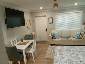 Un espace de vie compact comprenant un coin repas, un ensemble de canapés, un téléviseur fixé au mur et une fenêtre avec des stores.