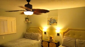 Quarto de hotel com duas camas, ventoinha no teto, luminárias fixadas na parede, uma mesinha com abajur e um quadro.