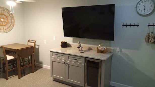 Quarto com televisão de tela plana fixada acima de uma bancada de cozinha, mesa de jantar com cadeiras e um relógio de parede.