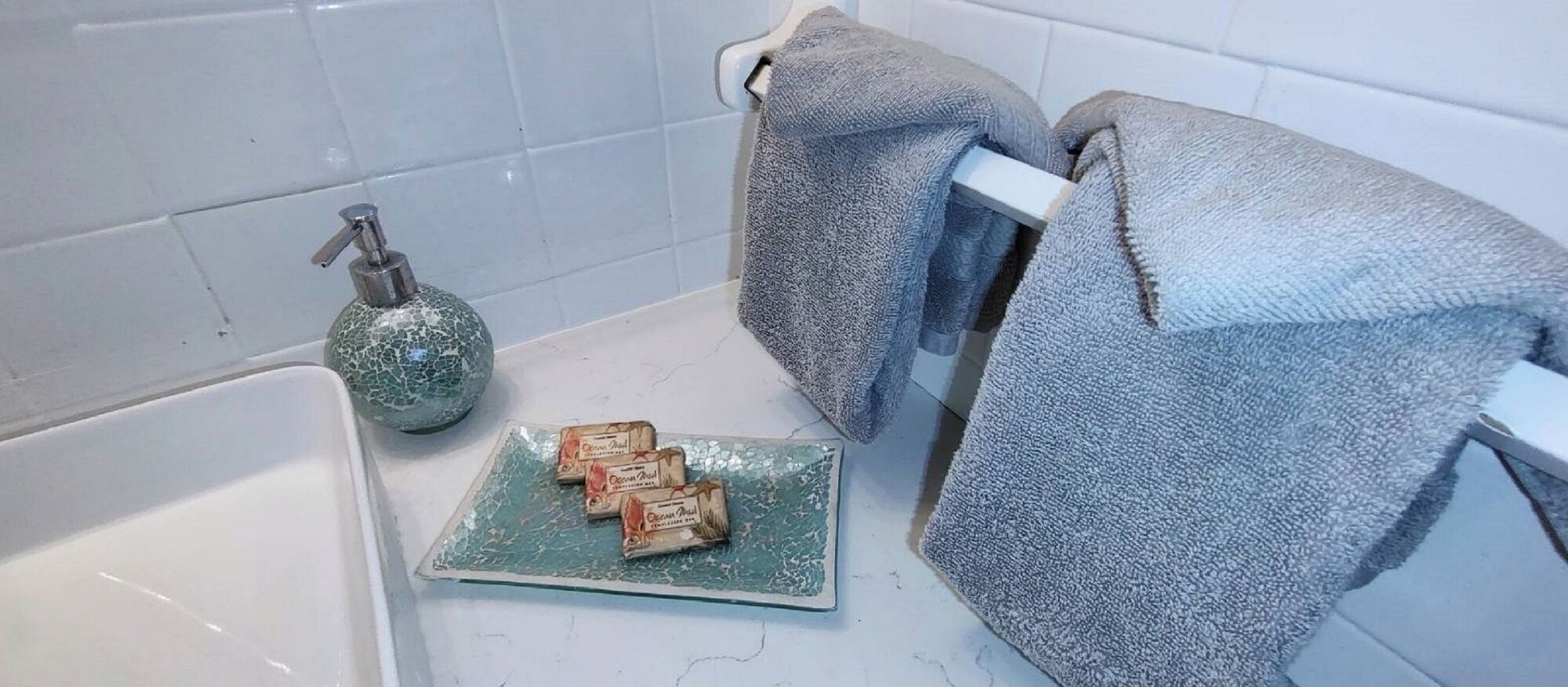 Ein Badezimmer mit einer Badewanne, einer Glastablett mit Seifenstücken, einem Seifenspender und zwei Handtüchern.