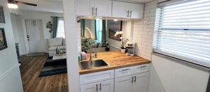 Een kleine keuken met witte kastjes, een houten aanrecht, een spoelbak en een raam met zonwering.