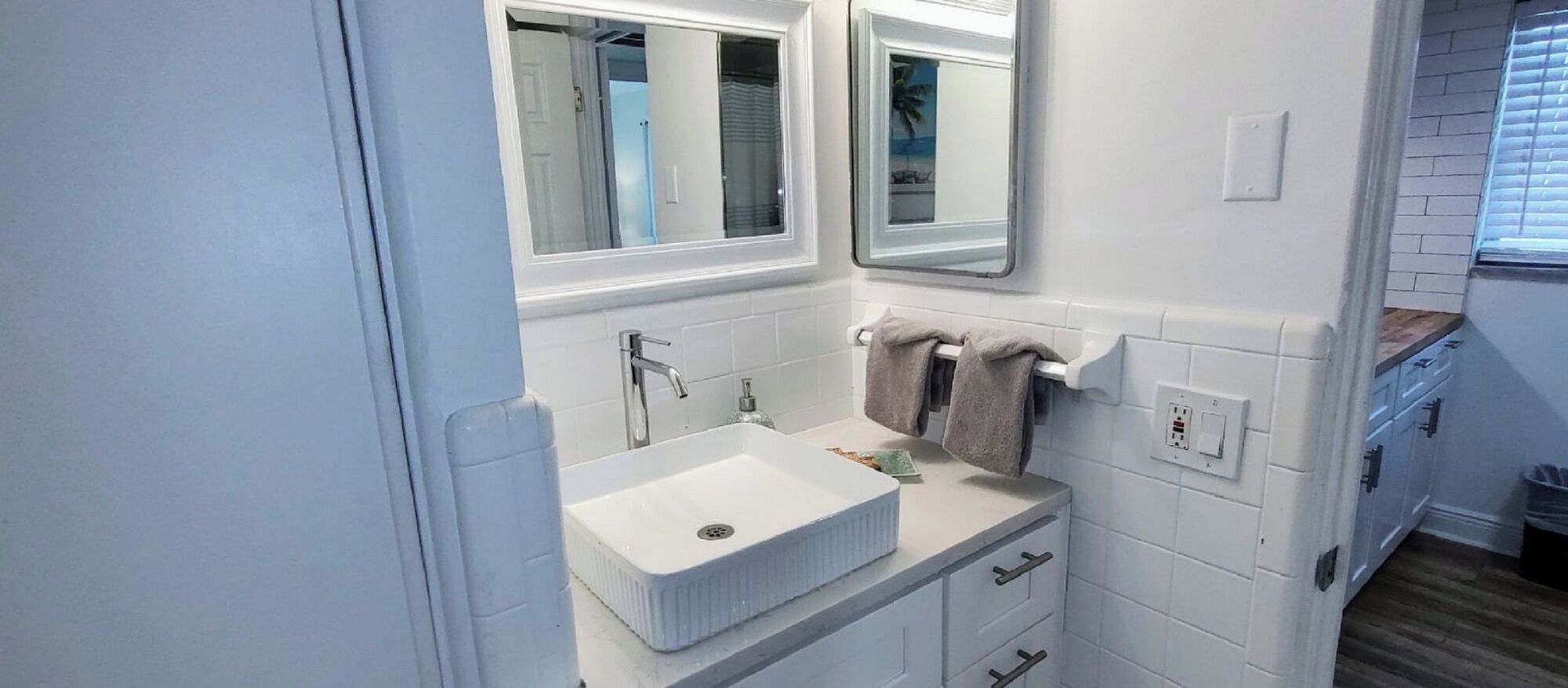 Ein Badezimmer mit einem weißen Waschbecken, zwei Spiegeln, einem Handtuchhalter und einem Fenster mit Jalousien.